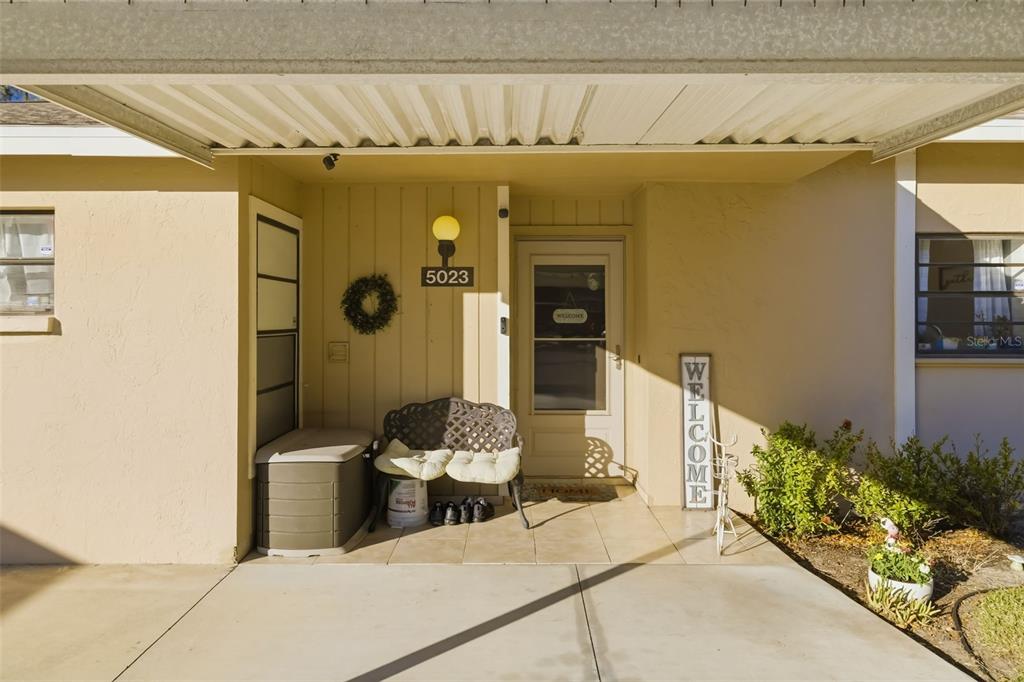 The exterior entrance features a covered concrete porch, a bench, and a welcome sign — 5023 VILLAGE GARDENS DRIVE UNIT 29, SARASOTA, FL