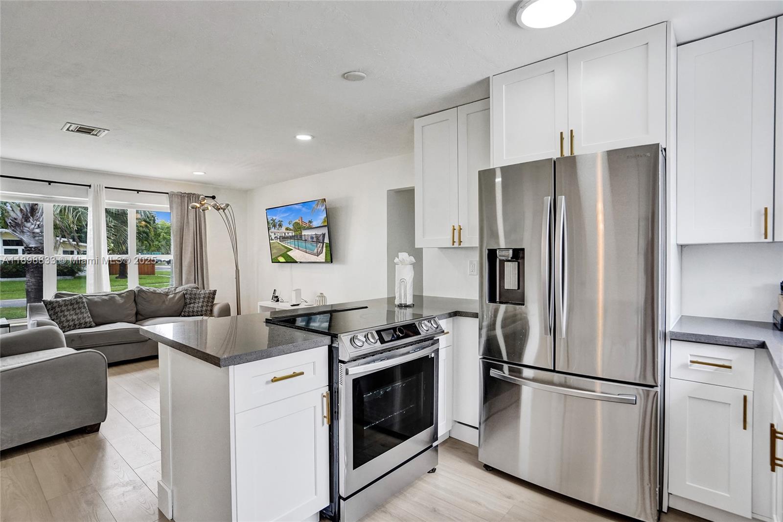 A kitchen with white cabinets and stainless steel appliances opens into a living room area — 1124 NE 6th St, Hallandale Beach FL 33009, HALLANDALE BEACH, FL