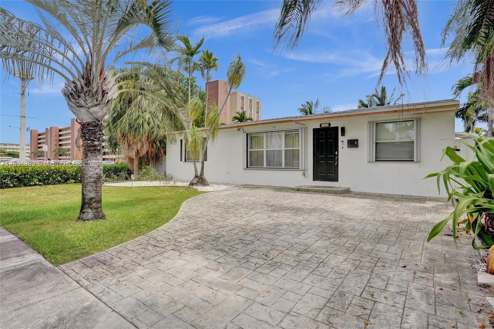 A single-story white house features a black front door and a stamped concrete driveway — 1124 NE 6th St, Hallandale Beach FL 33009, HALLANDALE BEACH, FL