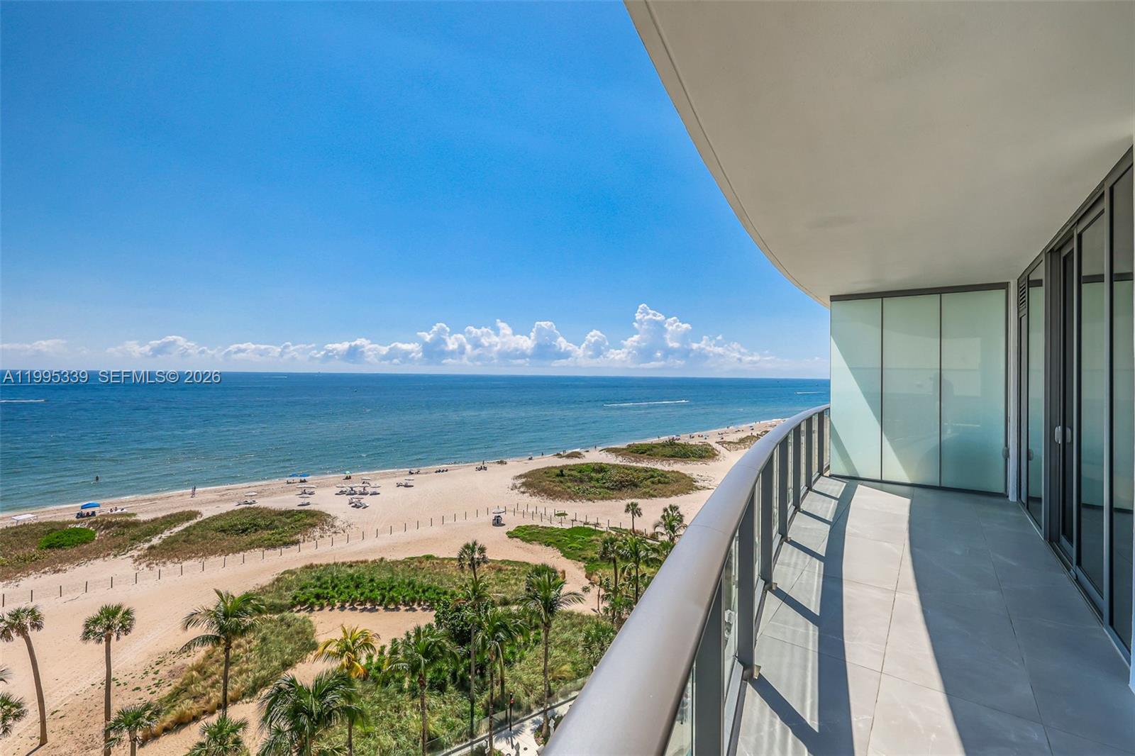 A high-rise balcony overlooks a sandy beach, ocean, and tropical landscaping under a clear sky — 1116 N Ocean Blvd # 806, Pompano Beach FL 33062, POMPANO BEACH, FL