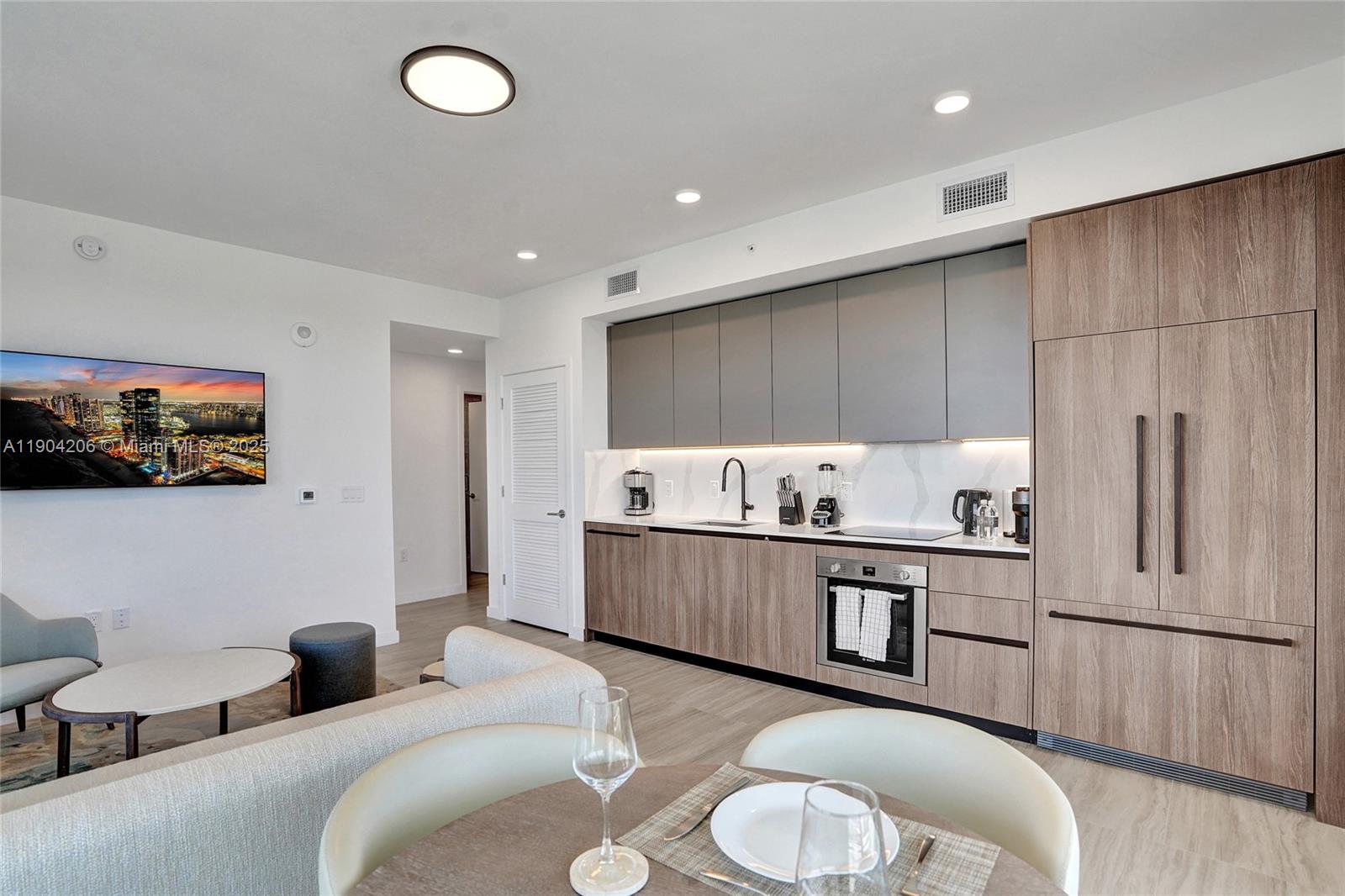 A modern kitchen with wood-toned cabinetry and grey upper cabinets opens into a living area — 580 72nd St # 810, Miami Beach FL 33141, MIAMI BEACH, FL