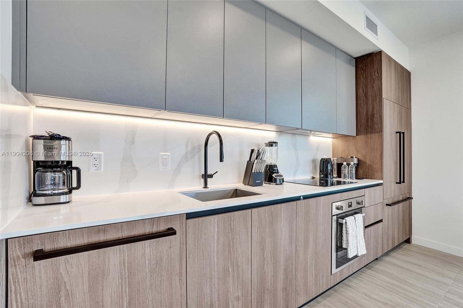 A modern kitchen features light wood cabinets, gray upper cabinets, white countertops, and stainless appliances — 580 72nd St # 810, Miami Beach FL 33141, MIAMI BEACH, FL