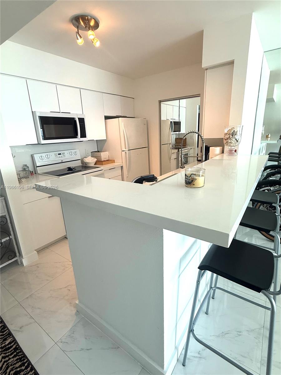 A white kitchen features a breakfast bar, stainless steel appliances, and light-colored tile flooring — 90 Alton Rd # 1106, Miami Beach FL 33139, MIAMI BEACH, FL