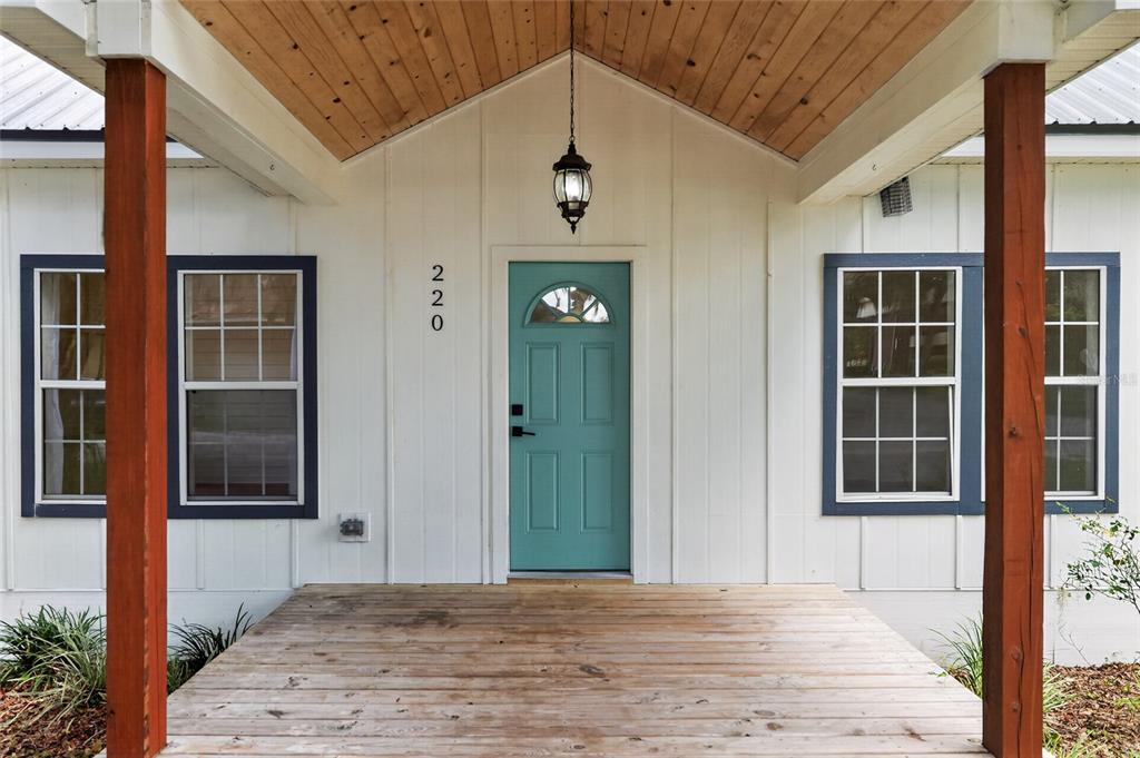 A covered wooden porch leads to a teal front door with a glass transom window — 220 NE 1ST AVENUE, WILLISTON, FL