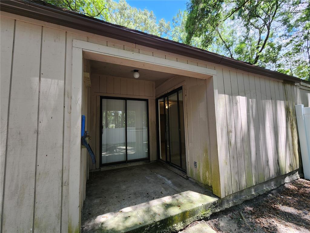 A concrete patio sits beneath a covered entryway with sliding glass doors and wood siding — 2018 SW 70TH TERRACE, GAINESVILLE, FL