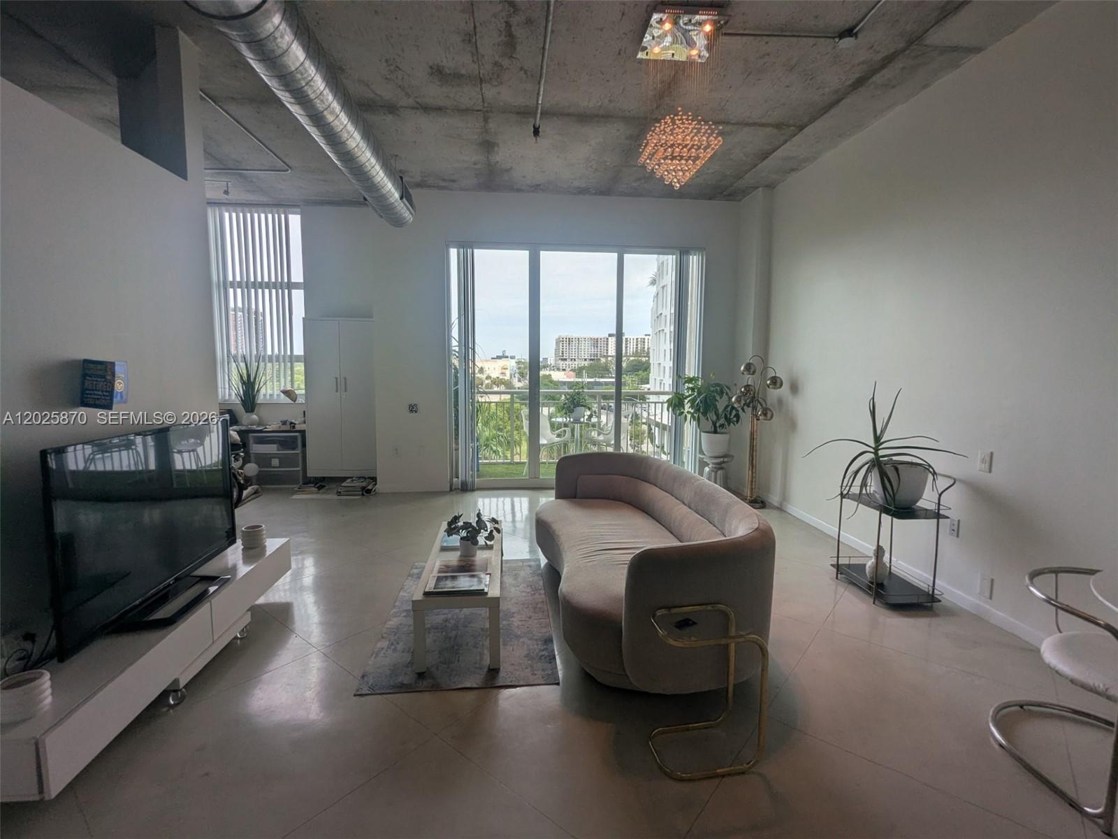 A modern living room features a curved sofa, concrete ceiling, exposed ductwork, and balcony access — 2001 Biscayne Blvd # 3506, Miami FL 33137, MIAMI, FL