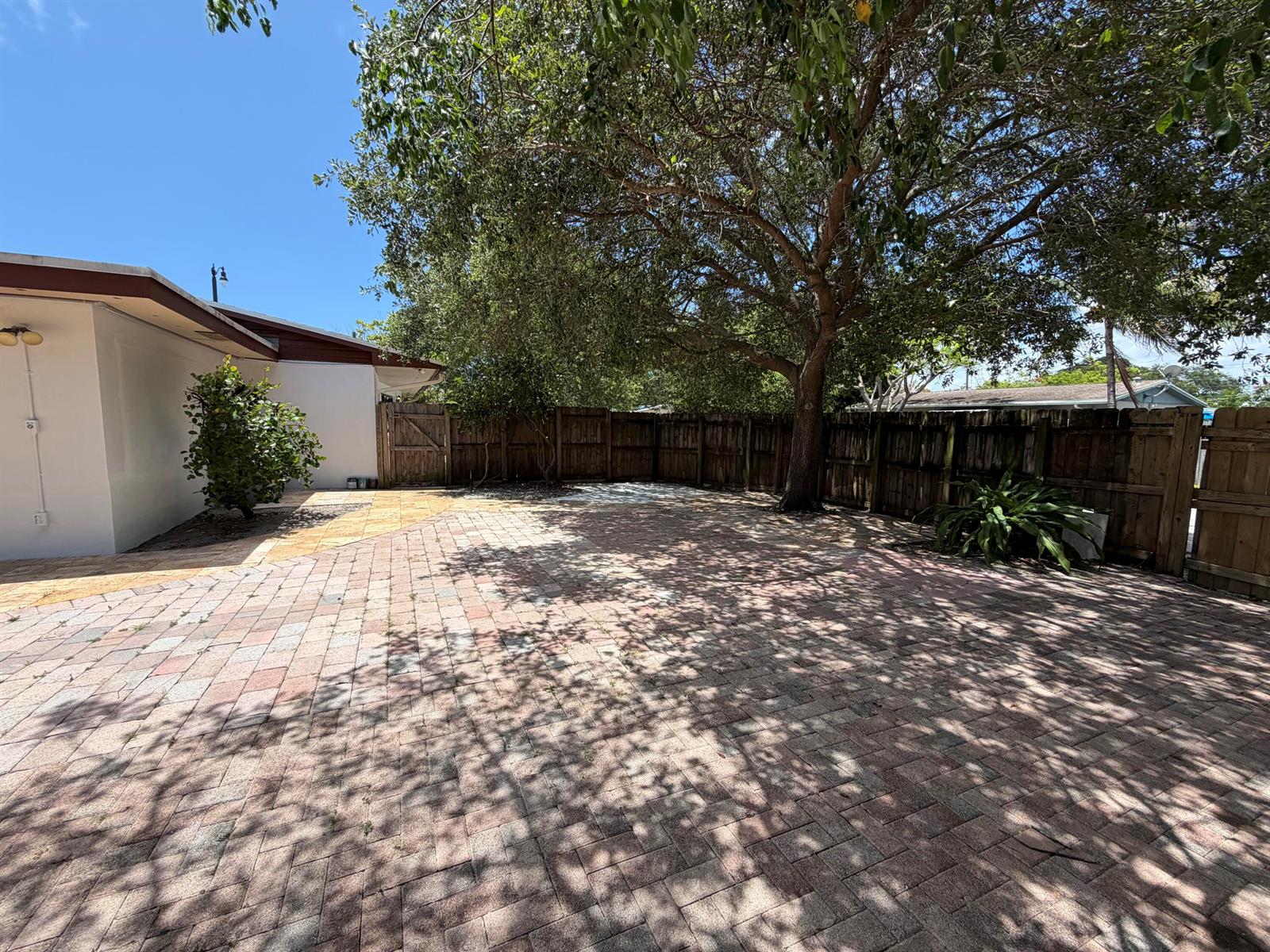 A paved patio area sits beside a house with a large tree and wooden fence — 4250 NE 12th Avenue, Pompano Beach FL 33064, POMPANO BEACH, FL