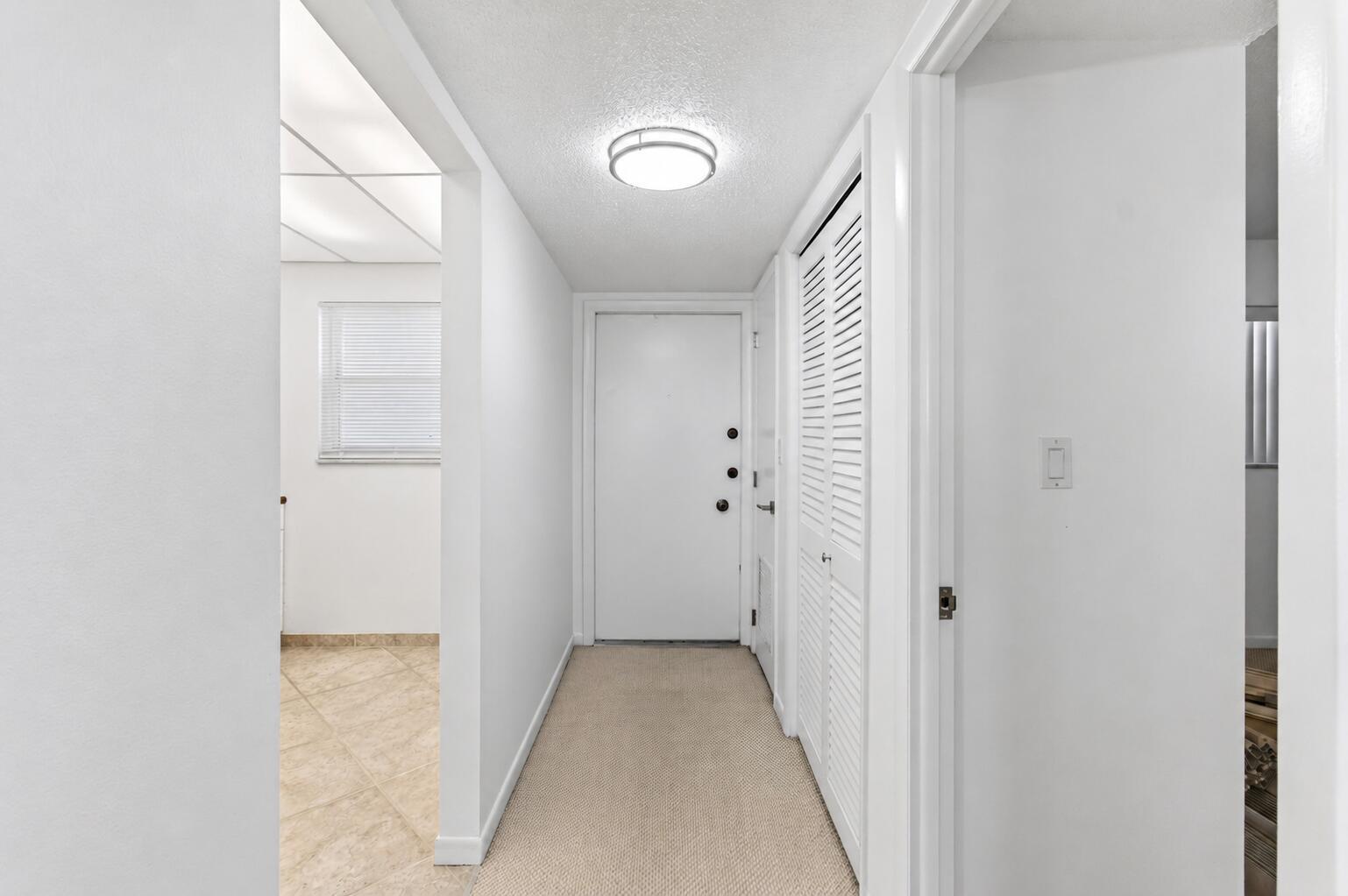 A hallway with carpeted flooring leads to a white front door and a louvered closet — 555 NW 4 Avenue # 4230, Boca Raton FL 33432, BOCA RATON, FL