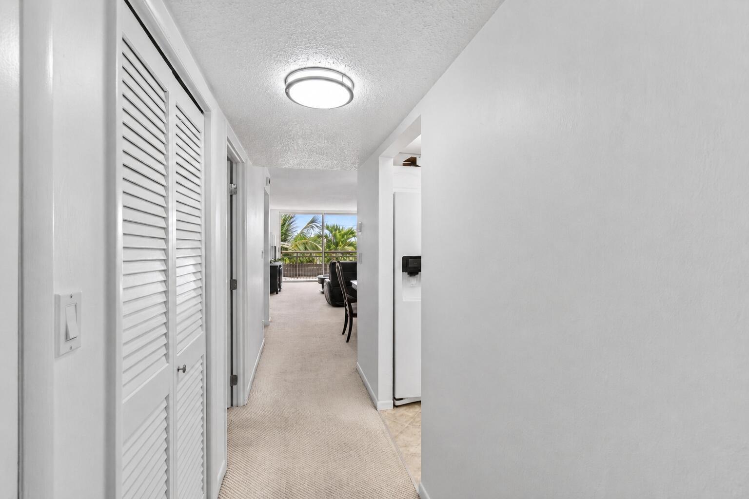 A hallway with white walls and carpet leads toward a living area with a balcony view — 555 NW 4 Avenue # 4230, Boca Raton FL 33432, BOCA RATON, FL