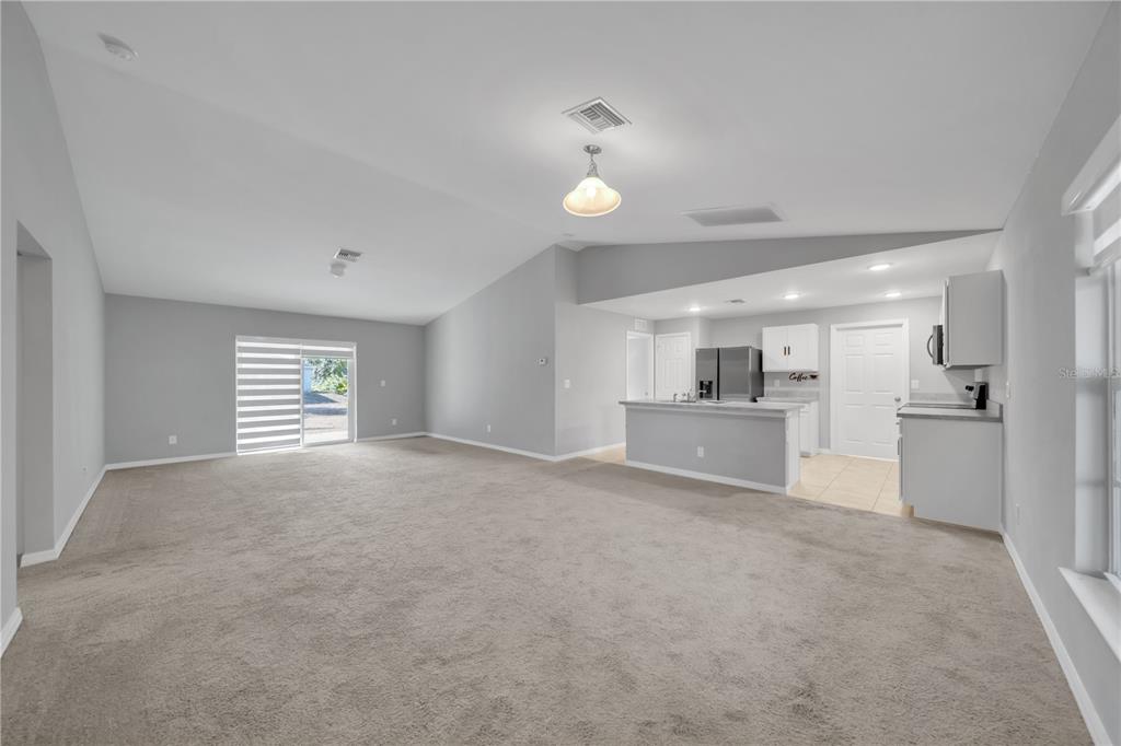 A spacious living room with beige carpeting flows into an open kitchen with white cabinets — 2617 53RD ST W, LEHIGH ACRES, FL