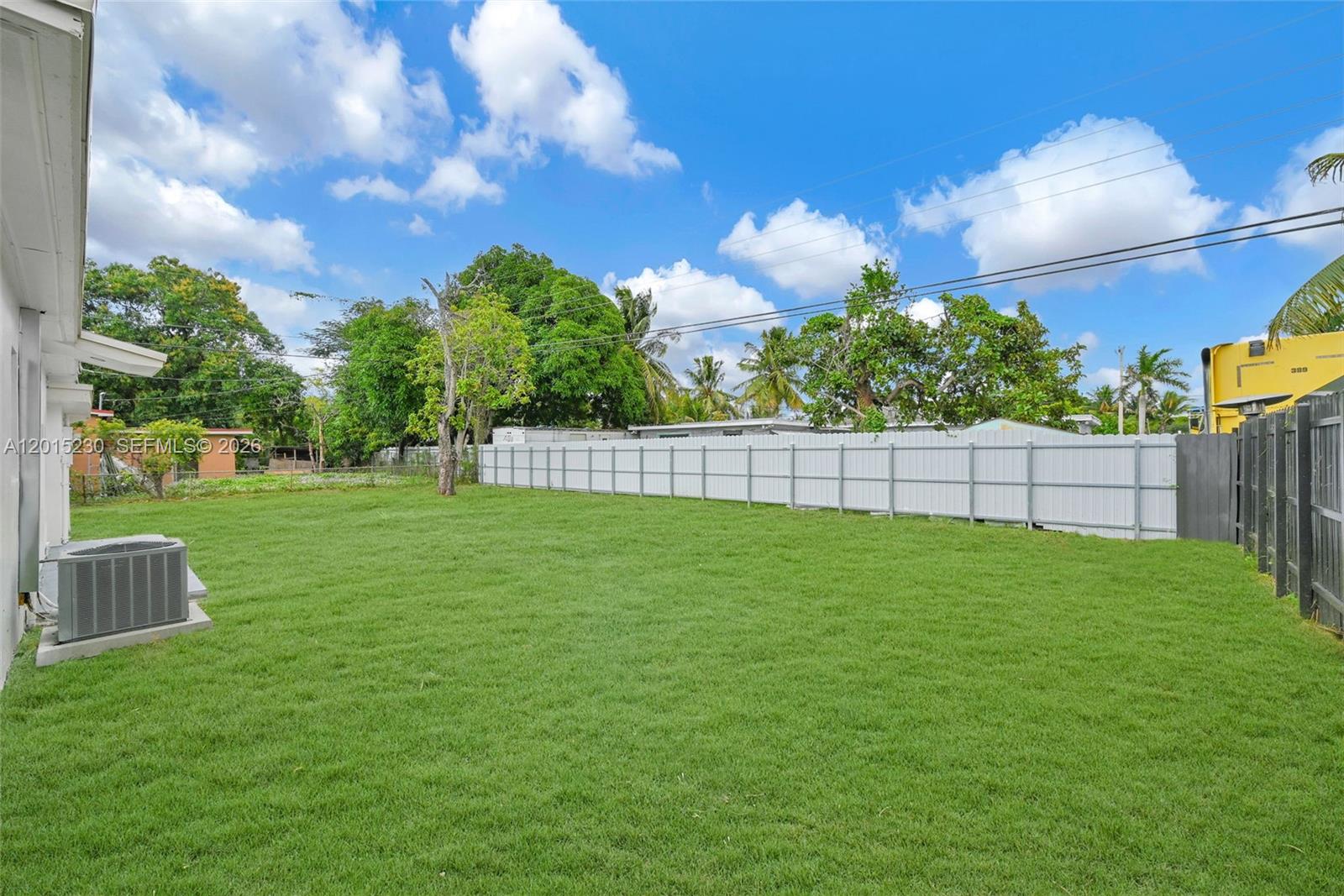 A grassy backyard features a white vinyl fence, an air conditioning unit, and mature trees — 12621 NW 22nd Ct, Miami FL 33167, MIAMI, FL
