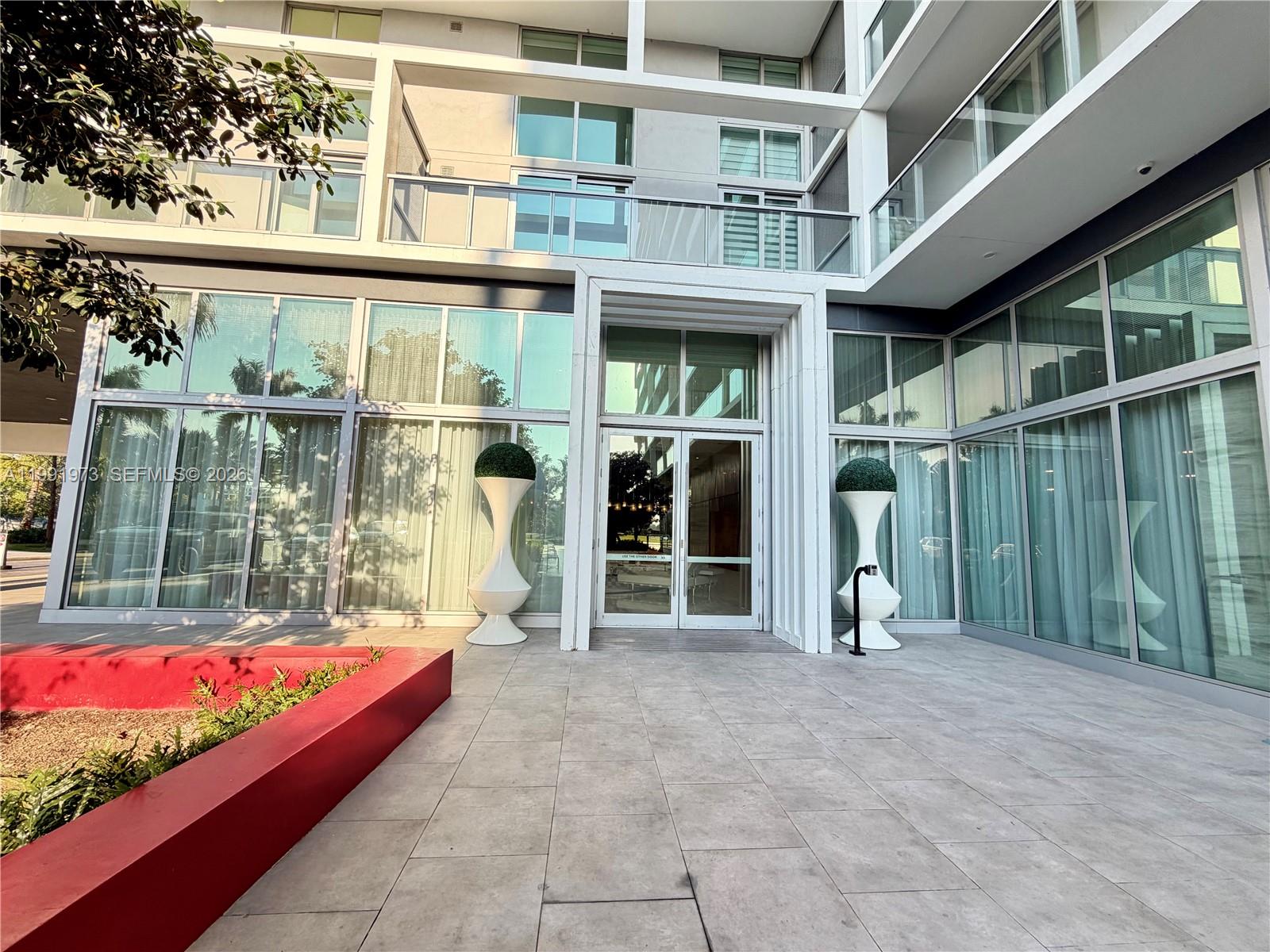 The exterior entrance of a modern building features glass walls, double doors, and decorative planters — 2000 metropica way # 2208, Sunrise FL 33323, SUNRISE, FL
