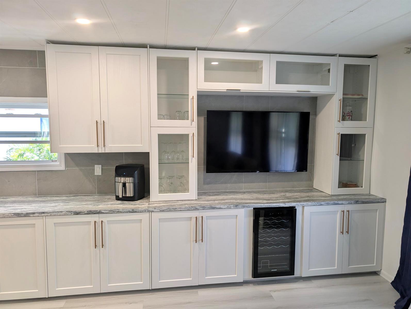 A modern kitchen features white cabinetry, gray stone countertops, a wall-mounted television, and a wine cooler — 37045 Exuma Bay, Boynton Beach FL 33436, BOYNTON BEACH, FL