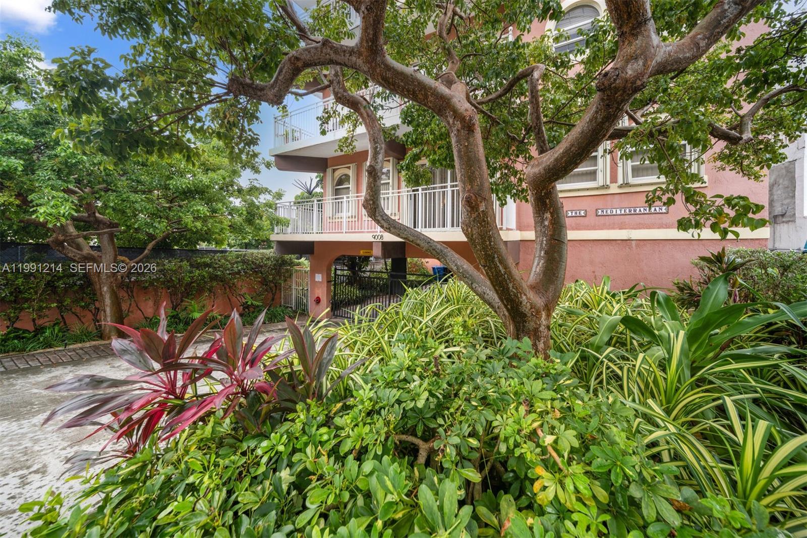 A multi-story pink building with balconies is partially obscured by trees and lush landscaping — 9008 Collins Ave # 402, Surfside FL 33154, SURFSIDE, FL