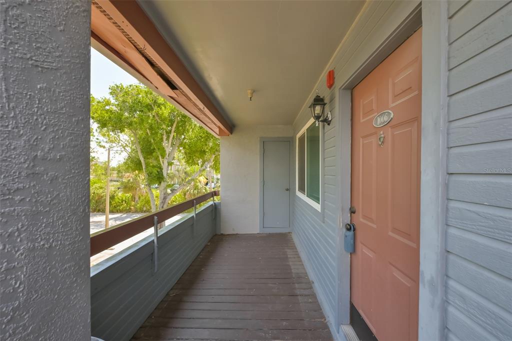 An exterior view of a covered apartment balcony with a wooden floor and peach-colored door — 10265 GANDY BOULEVARD N UNIT 1006, ST PETERSBURG, FL