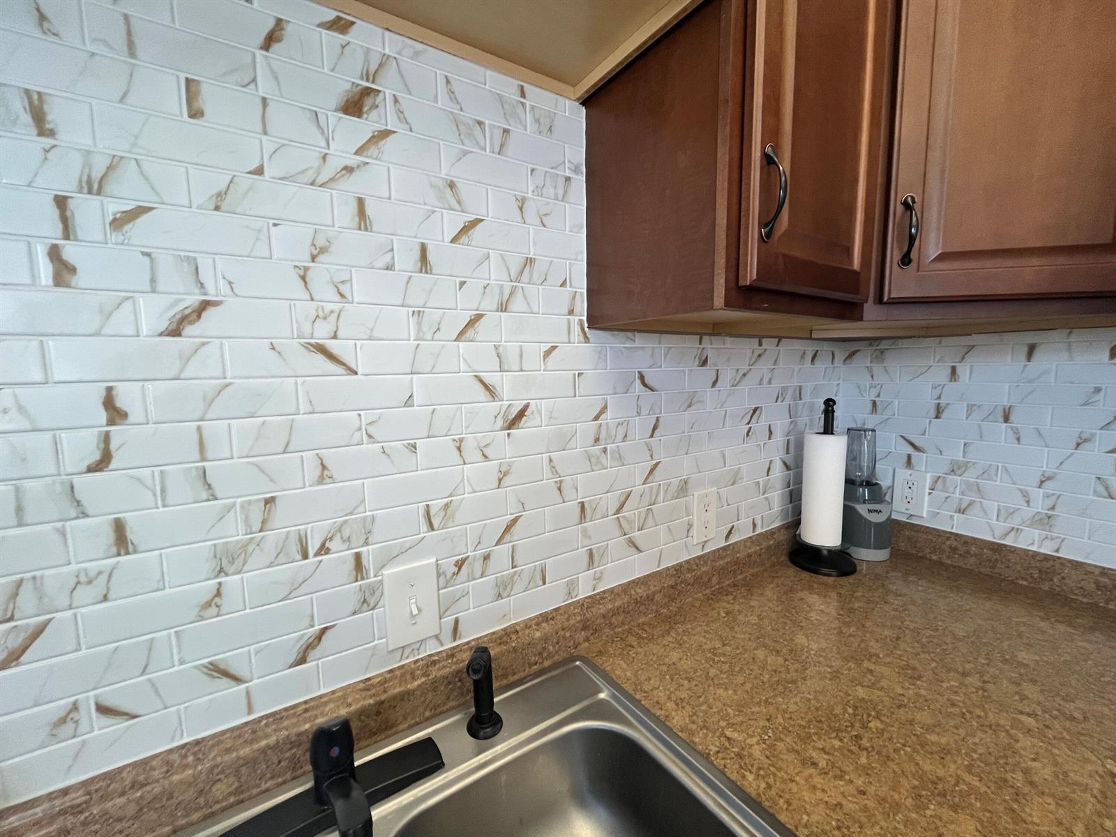 A kitchen sink area features a marble-look subway tile backsplash and brown wooden cabinets — 289 Brittany G, Delray Beach FL 33446, DELRAY BEACH, FL