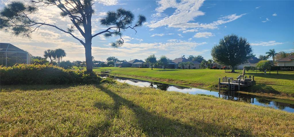 A grassy backyard features a canal with a small wooden dock under a sunny sky — 437 ROTONDA CIRCLE, ROTONDA WEST, FL