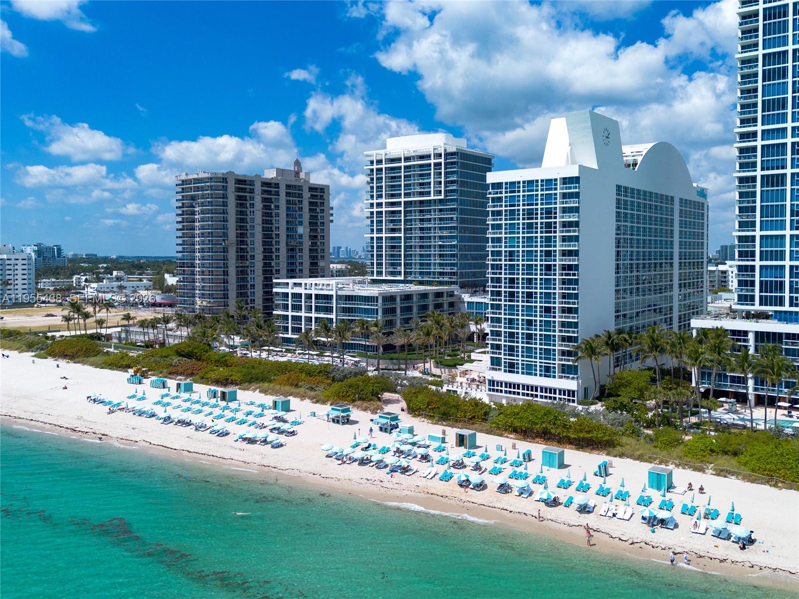 An aerial view of a sandy beach with lounge chairs in front of several high-rise buildings — 6799 Collins Ave # 401, Miami Beach FL 33141, MIAMI BEACH, FL
