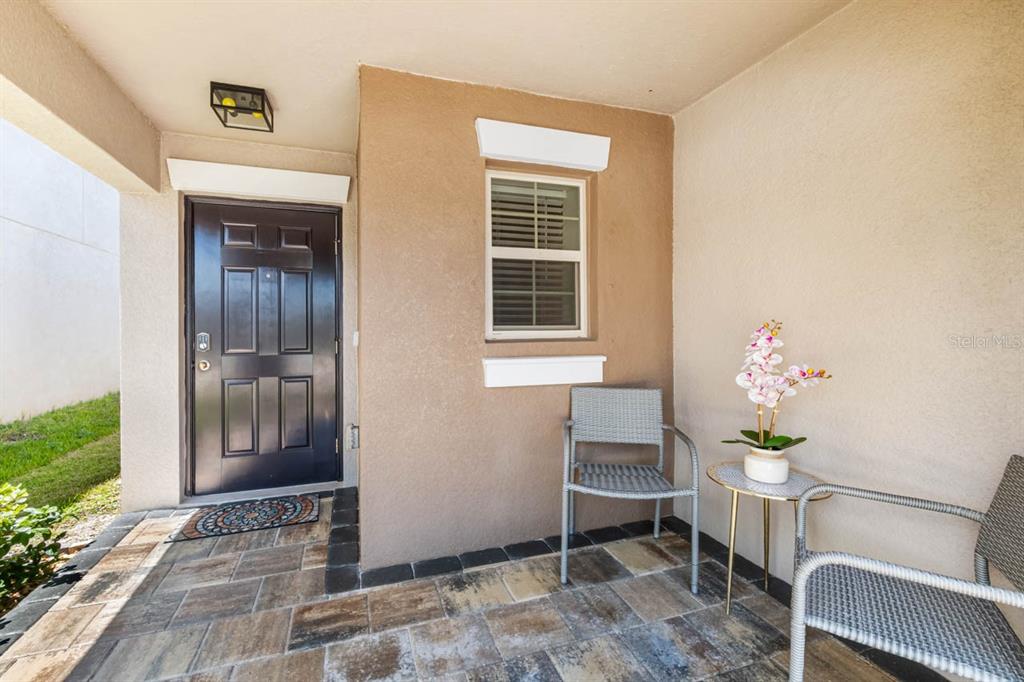 A covered entryway features a dark front door, a window, and two chairs on pavers — 15477 MILLER CREEK DRIVE, SUN CITY CENTER, FL