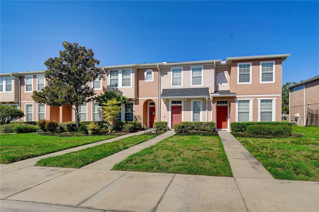 A row of two-story townhouses with tan stucco siding, red front doors, and concrete walkways — 6919 TOWERING SPRUCE DR, RIVERVIEW, FL