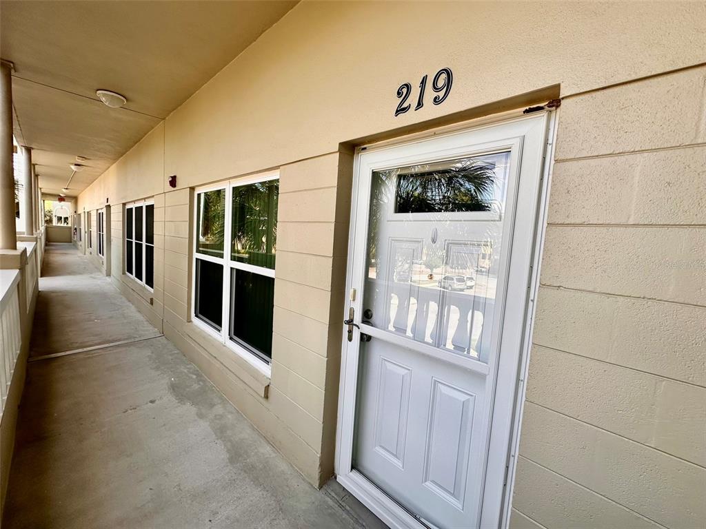 An exterior view of a tan building hallway featuring a white door labeled 219 — 2020 SHANGRILA DRIVE UNIT 219, CLEARWATER, FL