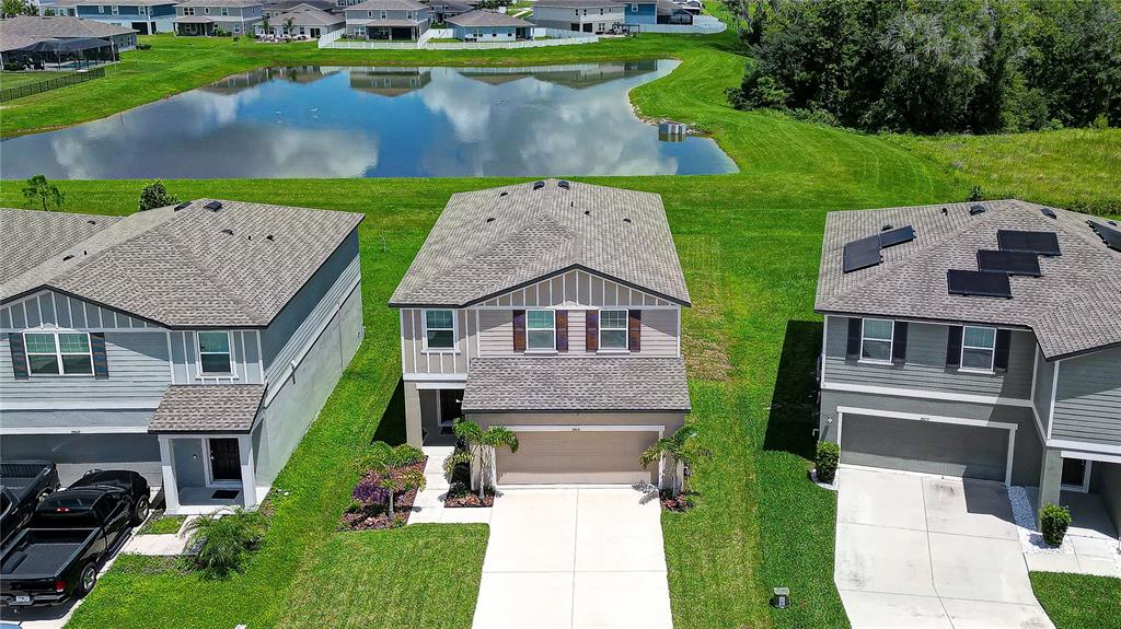 An aerial view shows three suburban houses with gray roofs situated near a retention pond — 35651 BUTTONWEED TRAIL, ZEPHYRHILLS, FL