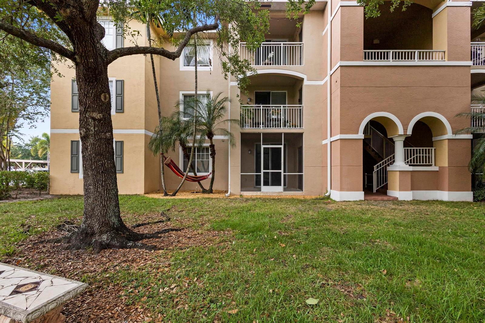 Exterior view of a multi-story apartment building with a screened patio and a hammock — 6450 Emerald Dunes Drive # 103, West Palm Beach FL 33411, WEST PALM BEACH, FL