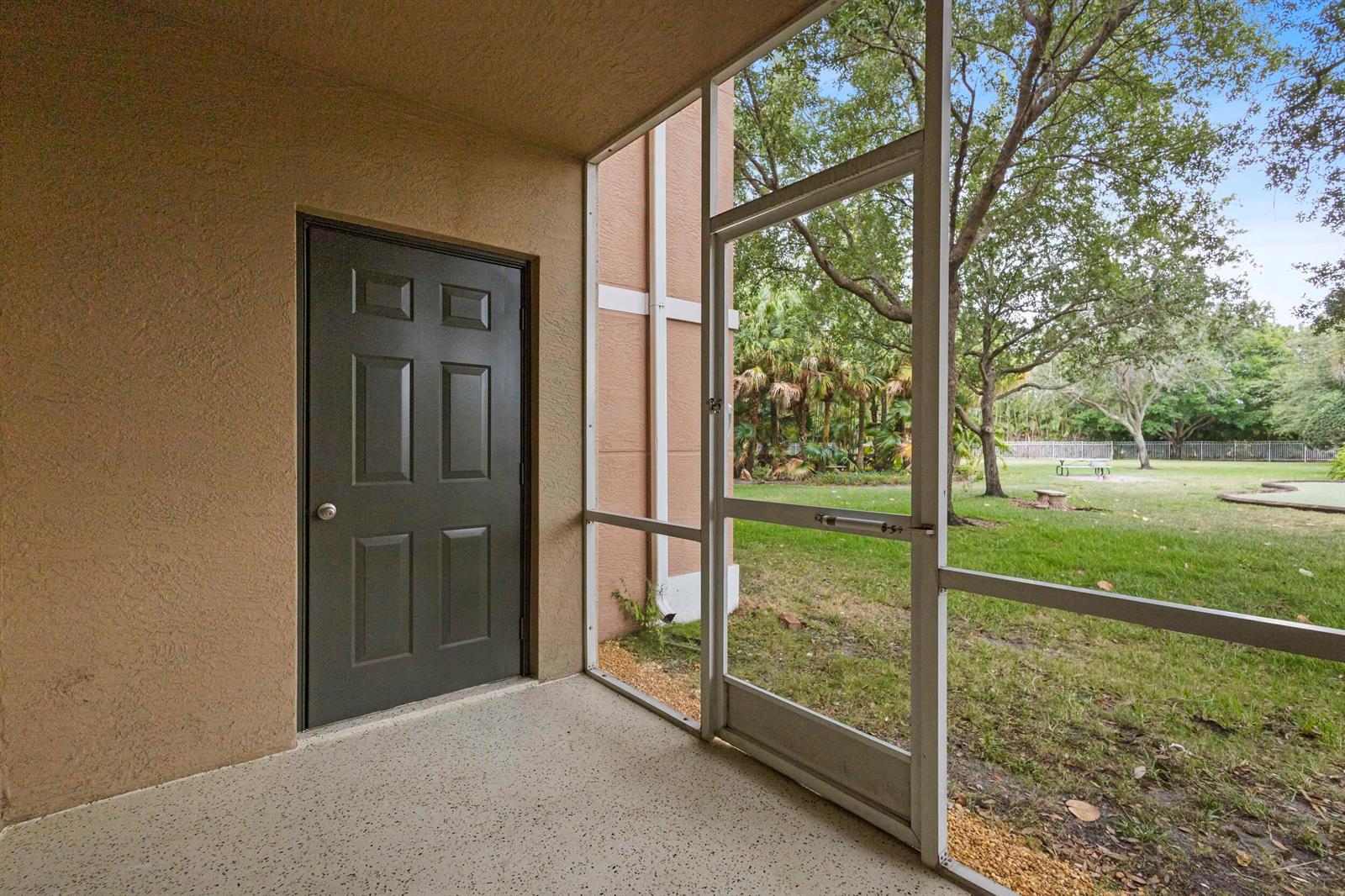 A screened-in patio with a dark door, concrete flooring, and a view of a lawn — 6450 Emerald Dunes Drive # 103, West Palm Beach FL 33411, WEST PALM BEACH, FL