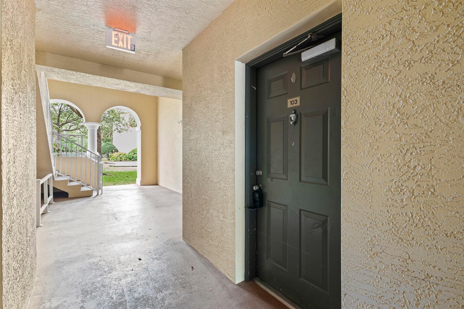 A dark brown apartment door labeled 103 sits in a beige stucco outdoor hallway — 6450 Emerald Dunes Drive # 103, West Palm Beach FL 33411, WEST PALM BEACH, FL
