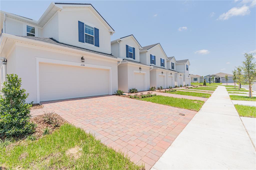 A row of white townhouses with brick driveways and blue shutters under a clear sky — 1174 SEASIDE MIST TRAIL, KISSIMMEE, FL