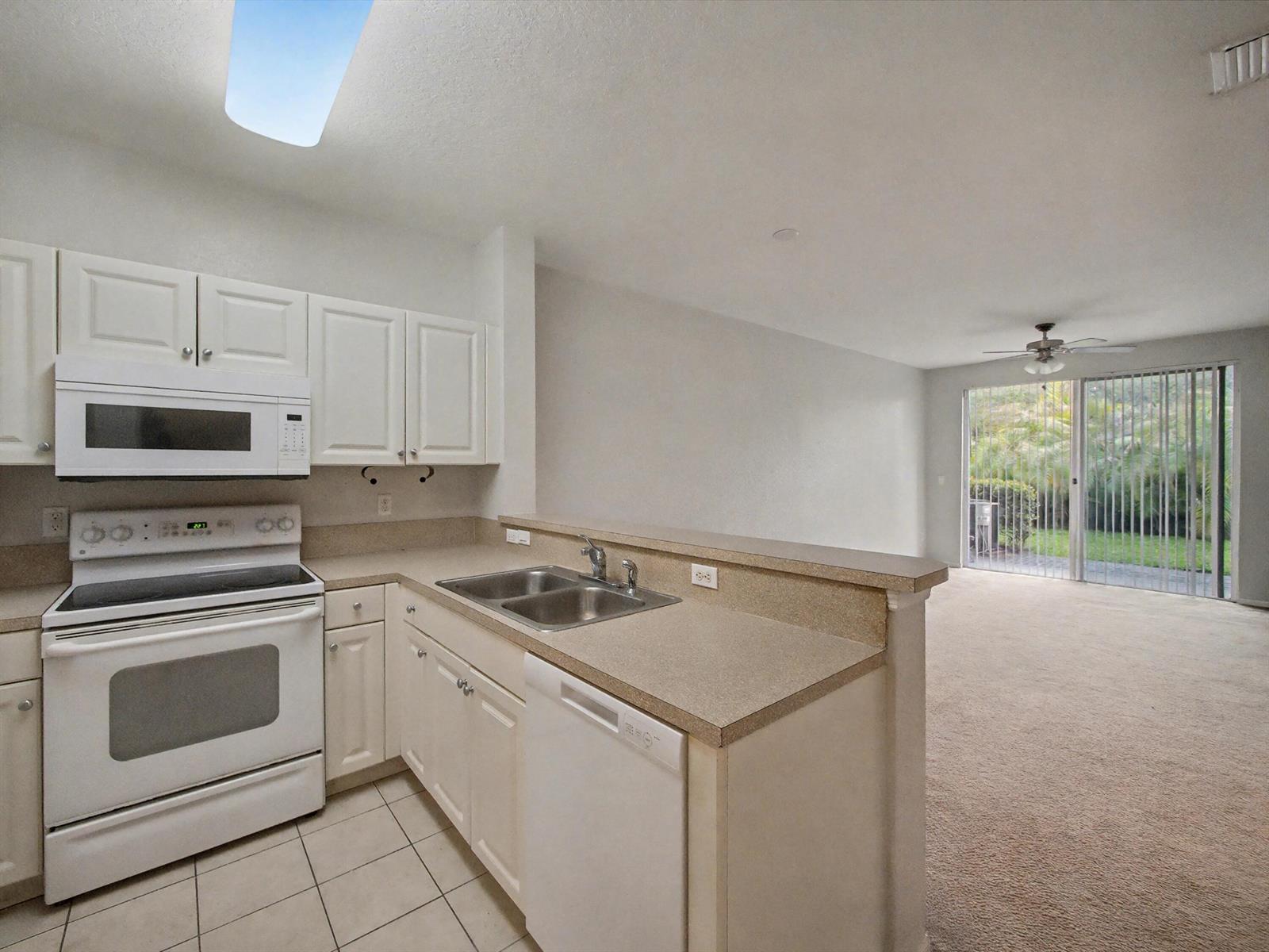 A kitchen with white cabinets and appliances overlooks a carpeted living area with sliding doors — 3425 Sonoma Drive, Riviera Beach FL 33404, RIVIERA BEACH, FL