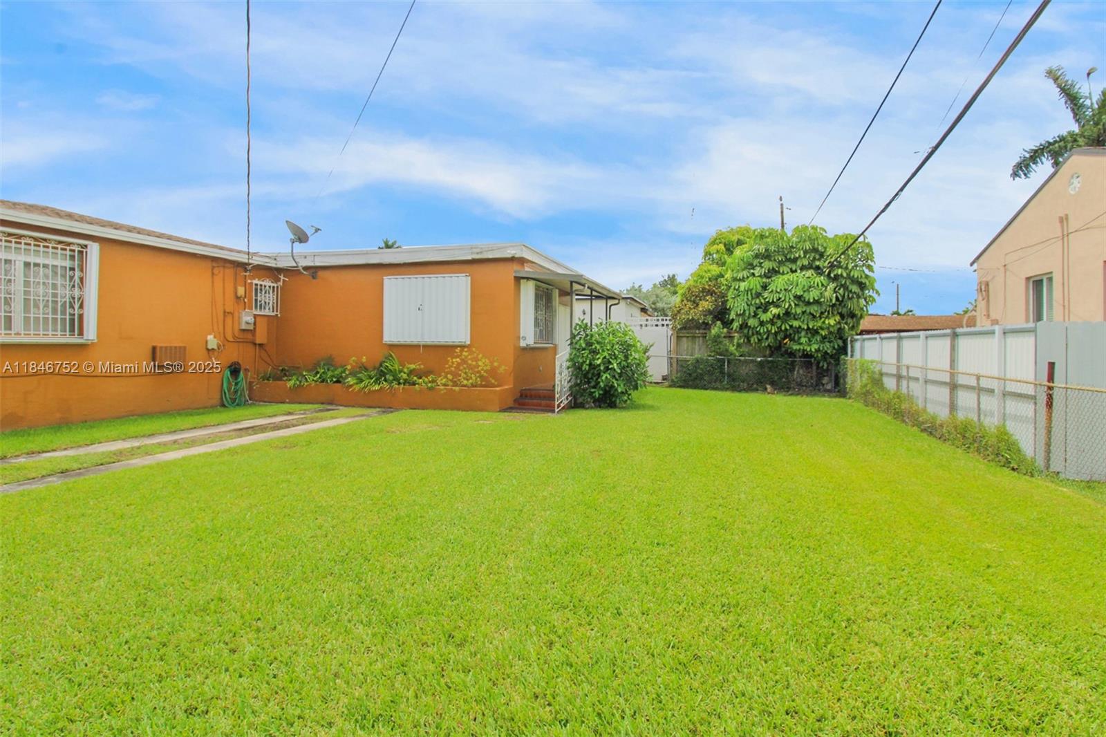 A grassy backyard features a single-story orange house with a concrete driveway and metal fencing — 3600 NW 19th Ter, Miami FL 33125, MIAMI, FL
