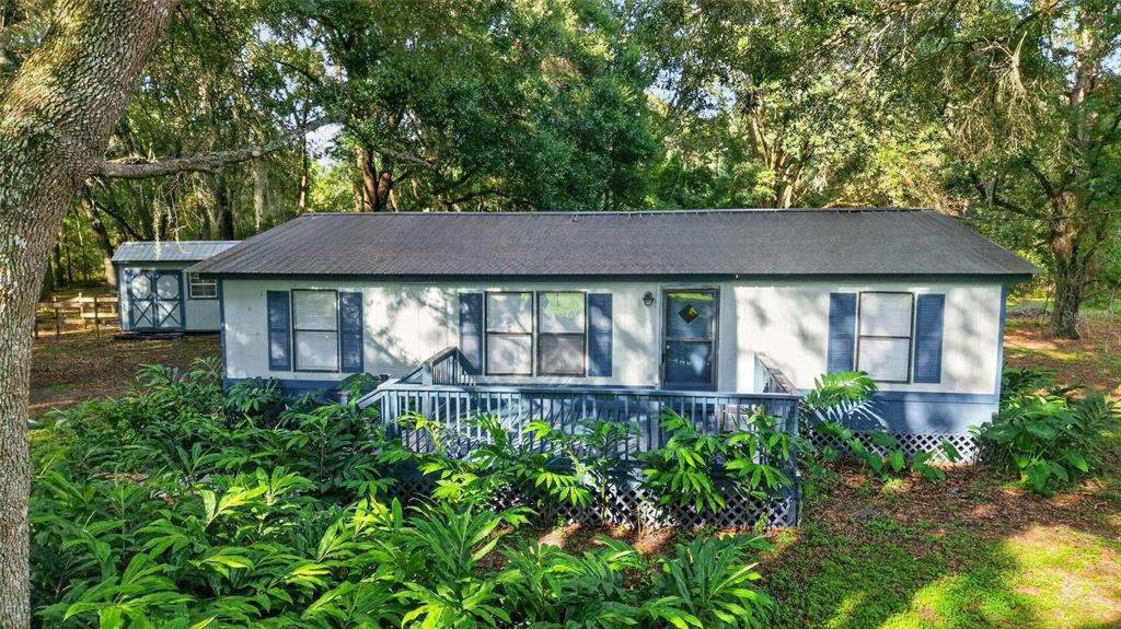 A single-story manufactured home with a front deck sits in a wooded, landscaped yard — 14210 NW HIGHWAY 464B, MORRISTON, FL