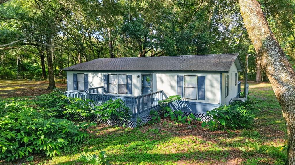A single-story manufactured home with a wooden deck sits in a wooded, grassy yard — 14210 NW HIGHWAY 464B, MORRISTON, FL