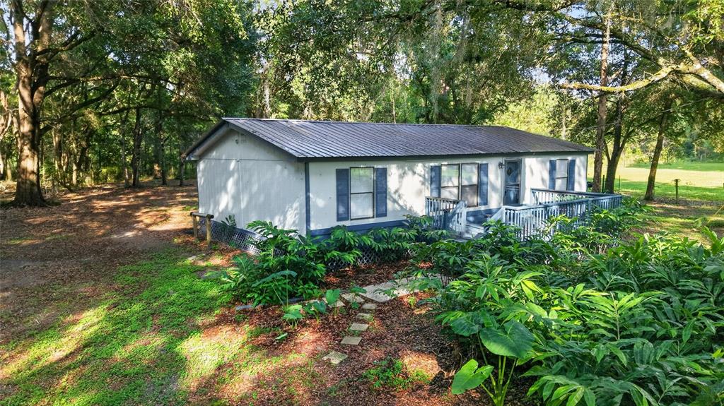 A single-story manufactured home with a metal roof sits in a wooded, grassy yard — 14210 NW HIGHWAY 464B, MORRISTON, FL