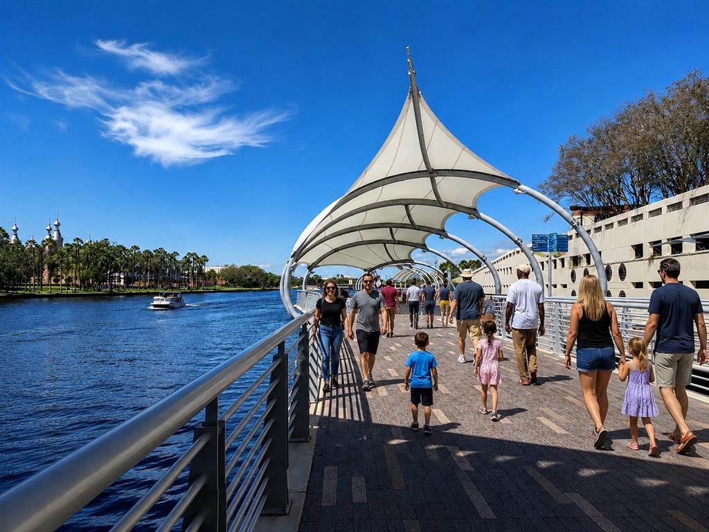 A group of people walks along a paved riverfront path under a white canopy structure — 1212 E 25TH AVENUE, TAMPA, FL