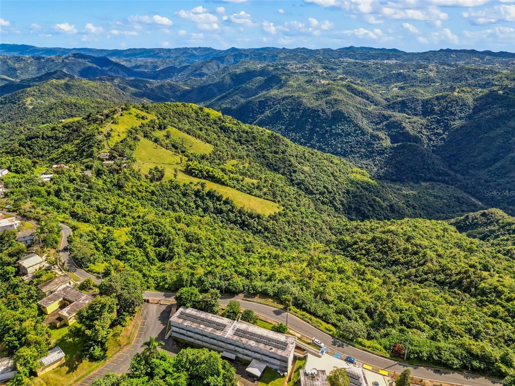 An aerial view shows a multi-story building situated along a road amidst lush, mountainous terrain — Carr 156 Km 20.1 INTERIOR BO HONDURAS, BARRANQUITAS, PR
