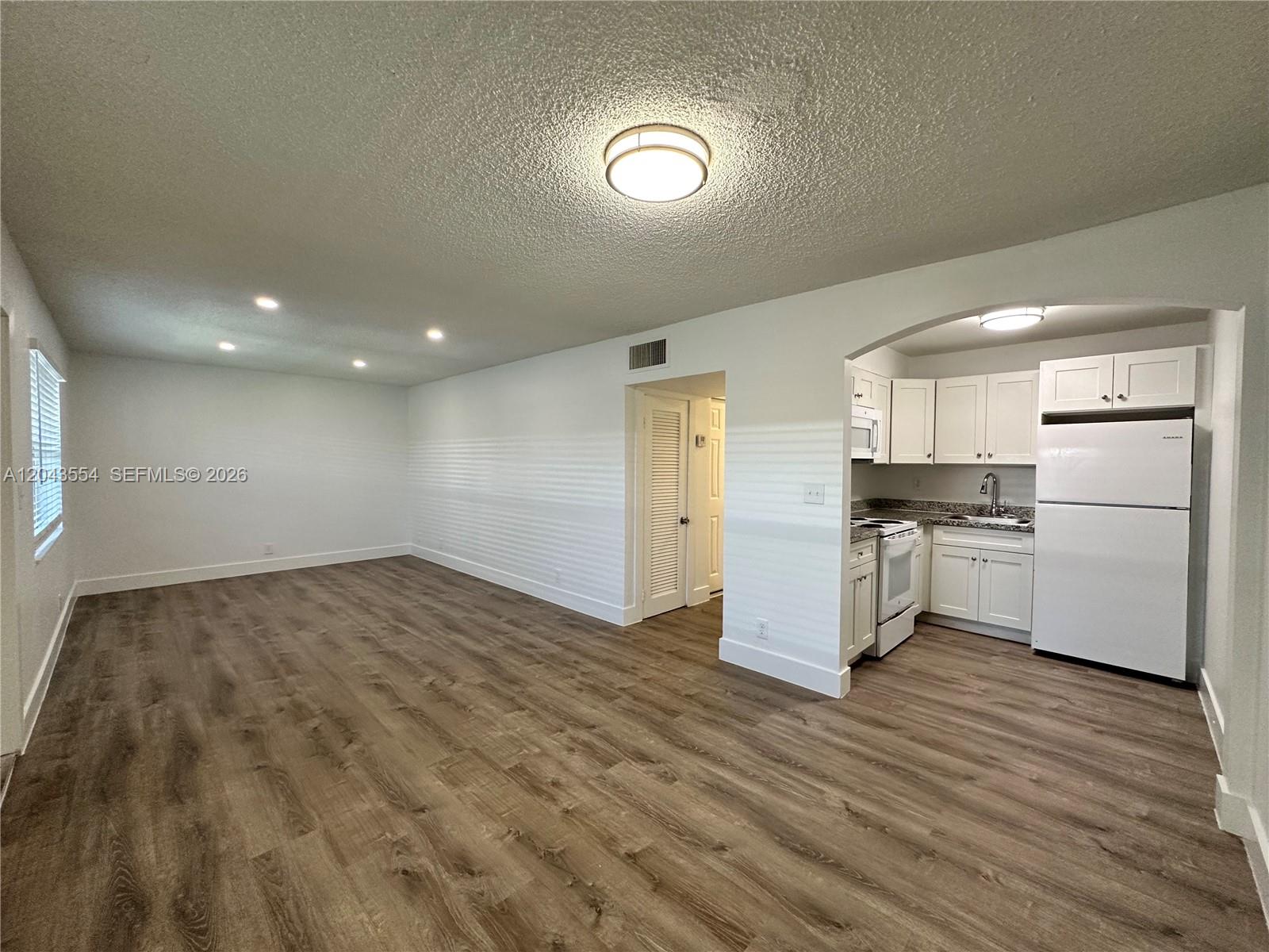 An open-concept living area with wood-look flooring leads into a small white kitchen — 2500 Johnson St # 19, Hollywood FL 33020, HOLLYWOOD, FL