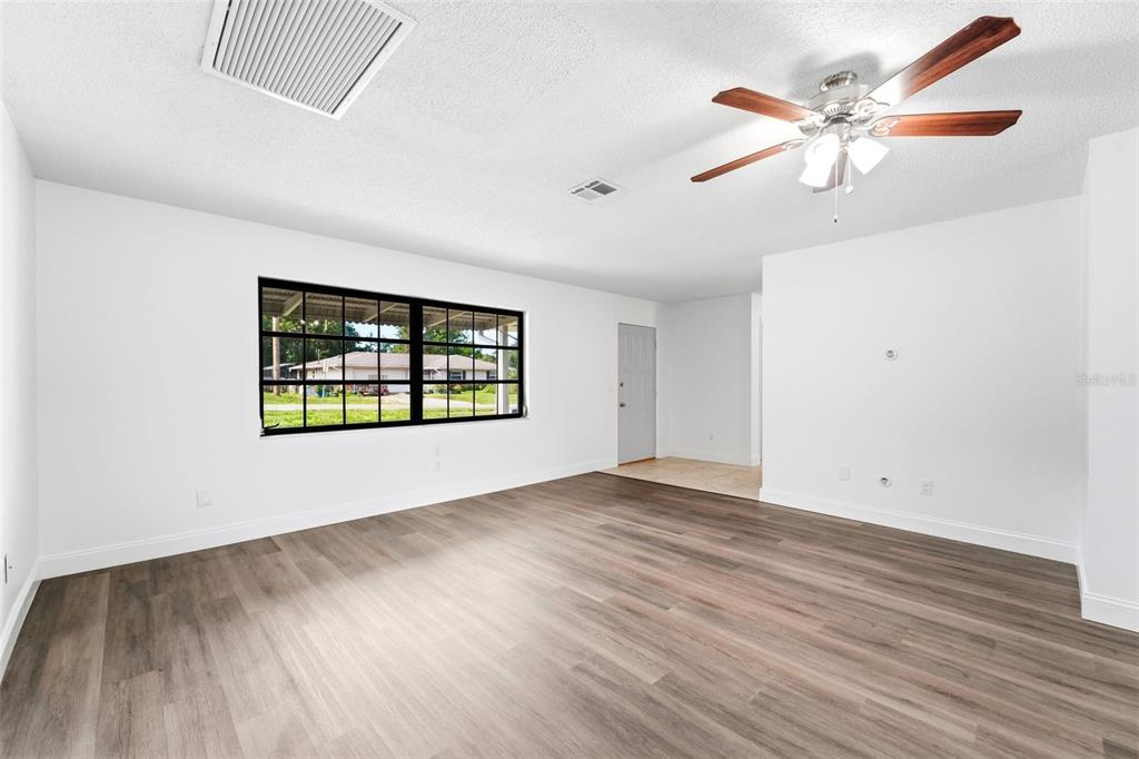 A bright, empty living room features wood-look flooring, a ceiling fan, and a large window — 1360 N OLD MILL DRIVE, DELTONA, FL