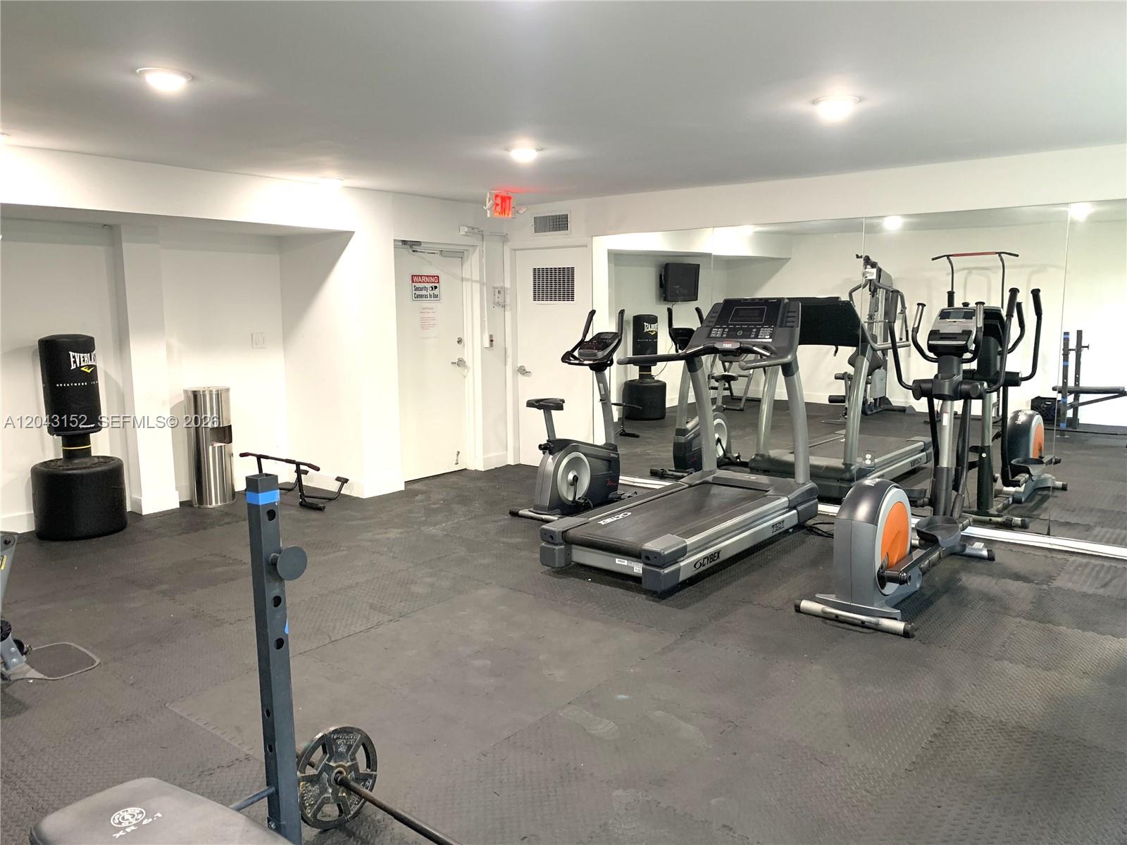 A gym room features rubber flooring, various exercise machines, a weight rack, and mirrored walls — SUNNY ISLES BEACH, FL