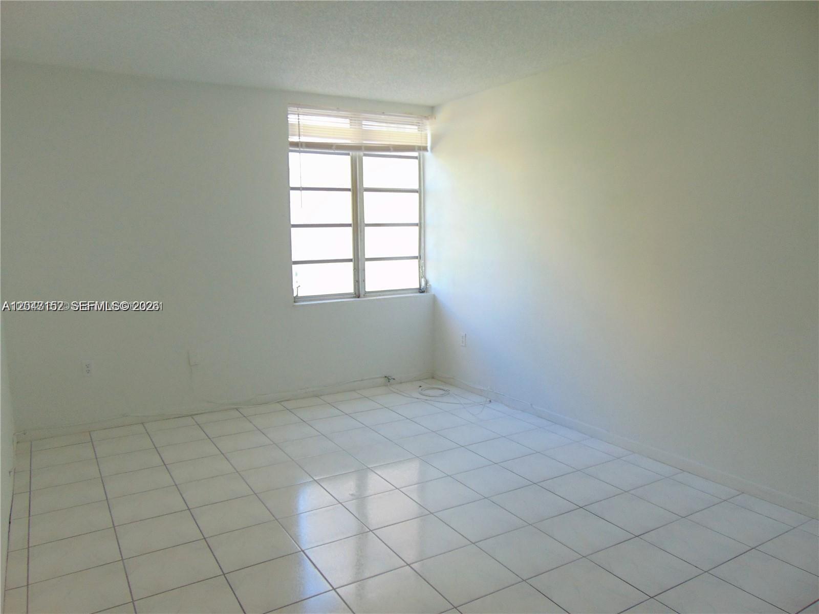 An empty room features white walls, white tile flooring, and a window with blinds — SUNNY ISLES BEACH, FL