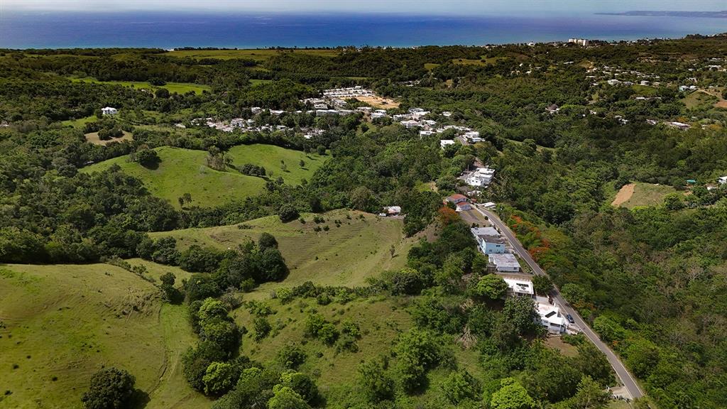 An aerial view shows a hillside road with houses overlooking a green landscape and the ocean — Km 1.1 CARR 400, RINCON, PR