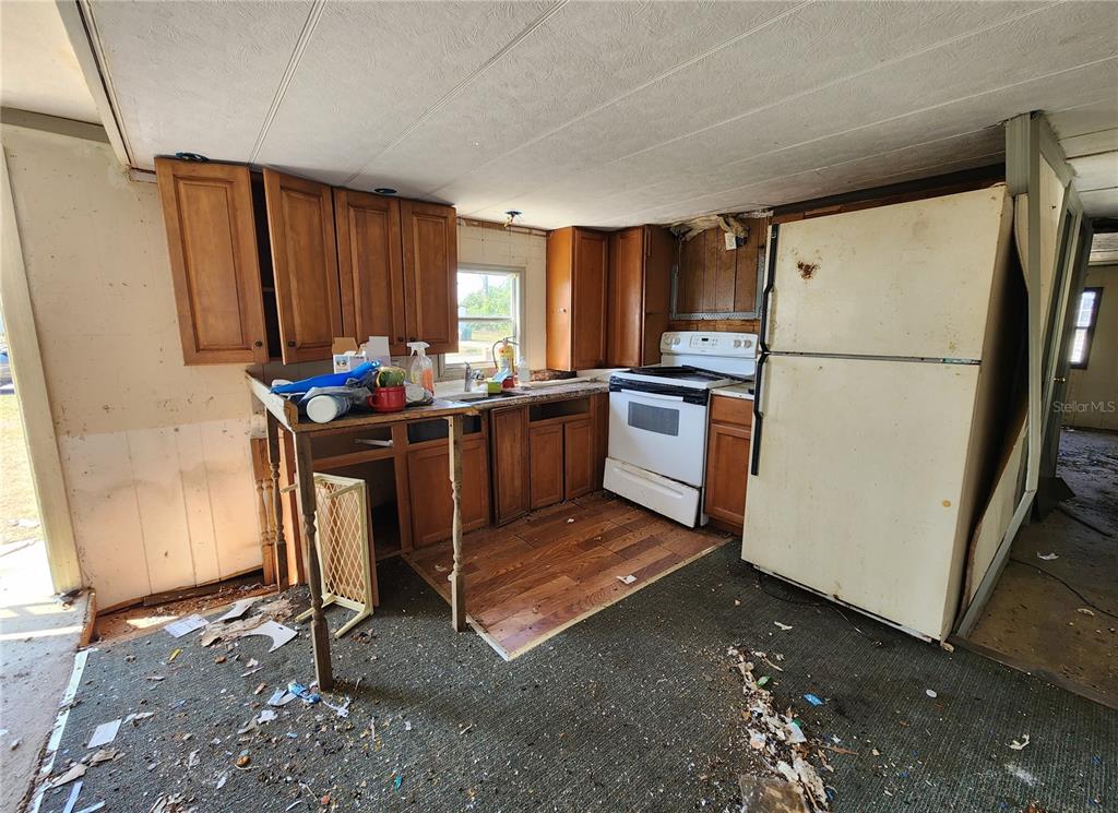 A dilapidated kitchen features wood cabinets, a white stove, a refrigerator, and debris-covered flooring — 3457 GAINSBORO ROAD, PORT CHARLOTTE, FL