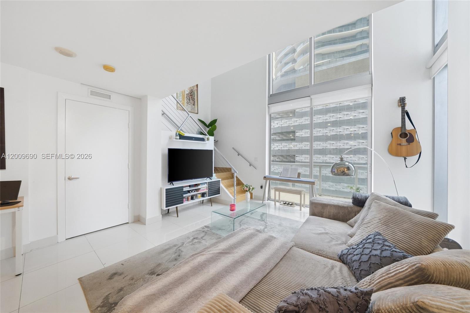A modern living room features a high ceiling, floor-to-ceiling windows, and a staircase — 1050 Brickell Ave # 1002, Miami FL 33131, MIAMI, FL