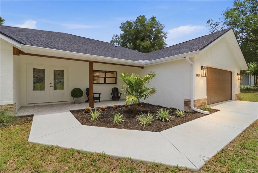 A white house exterior features a covered porch, a brown garage door, and landscaping — 1302 N MEDITERRANEAN WAY, INVERNESS, FL