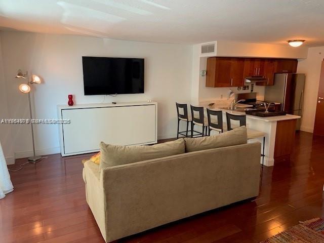A living room with a sofa, wall-mounted television, and an adjacent kitchen with wooden cabinets — 9499 Collins Ave # 205, Surfside FL 33154, SURFSIDE, FL