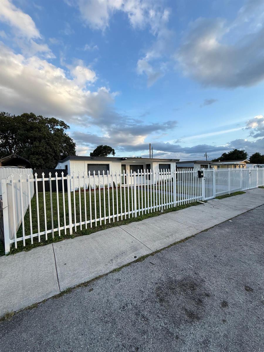 A white single-story house with a flat roof is enclosed by a tall metal fence — 1890 Rutland Street, Opa-Locka FL 33054, OPA-LOCKA, FL