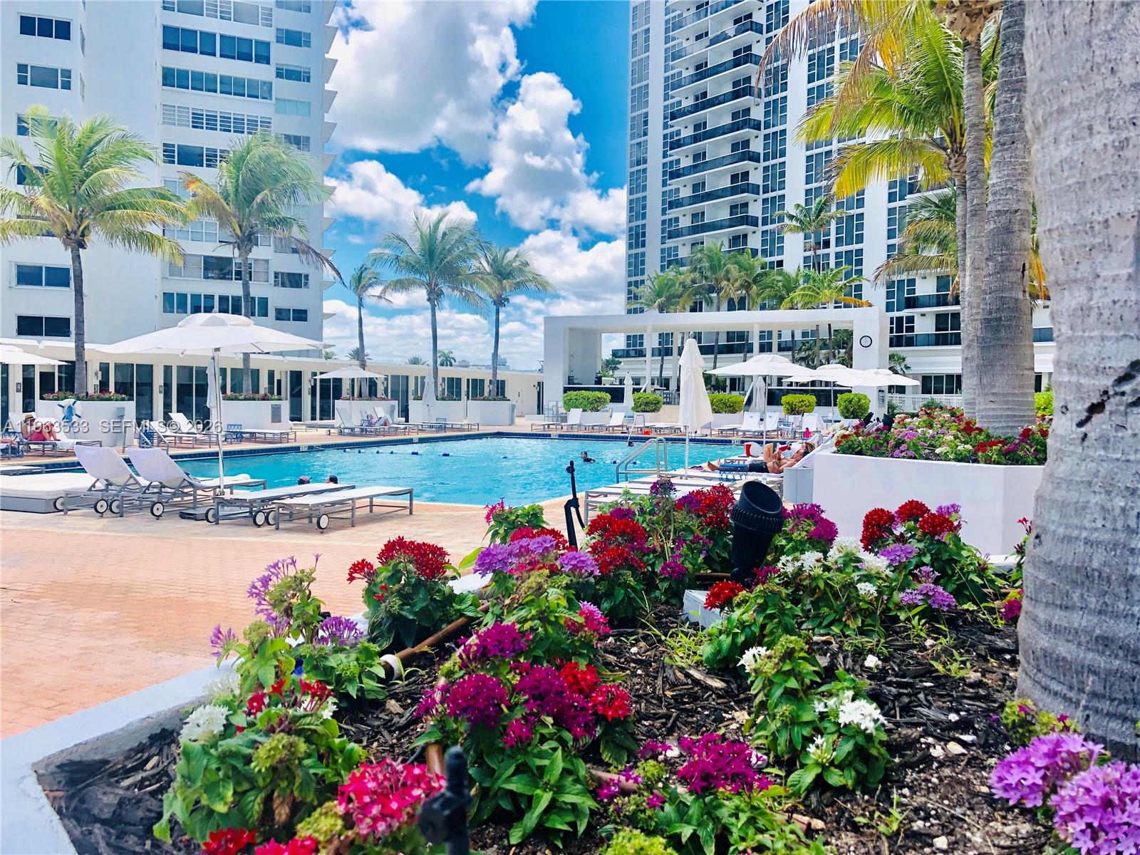 A swimming pool area with lounge chairs, palm trees, and flower beds between two tall buildings — 10275 Collins Ave # 427, Bal Harbour FL 33154, BAL HARBOUR, FL