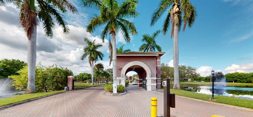 A gated entrance with palm trees, a brick driveway, and a pond under a blue sky — 10265 GANDY BOULEVARD N UNIT 1302, ST PETERSBURG, FL