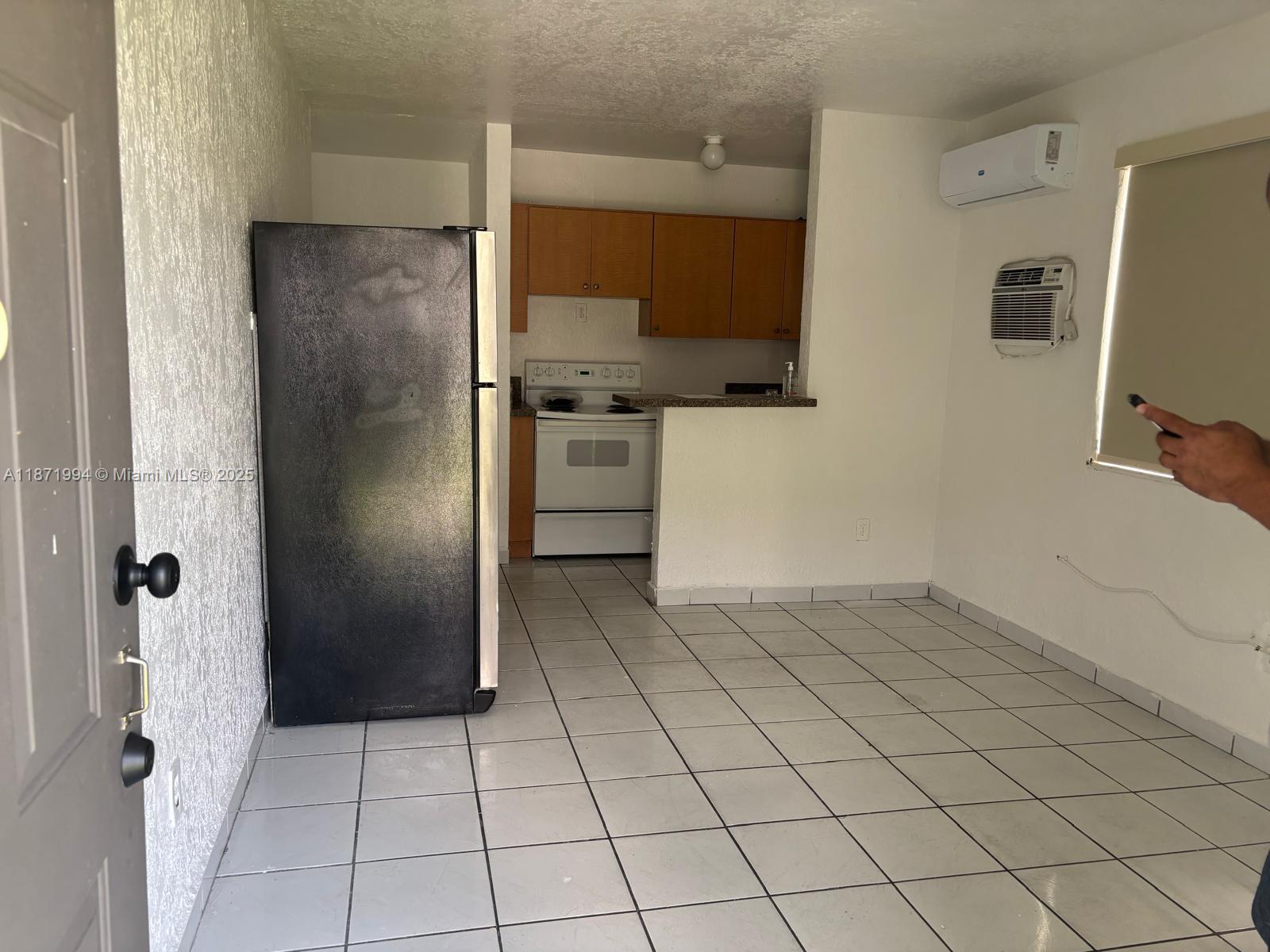 This small kitchen and living area features white tile flooring, a black refrigerator, and wood cabinets — 5100 NW 23rd Ave # 1, Miami FL 33142, MIAMI, FL